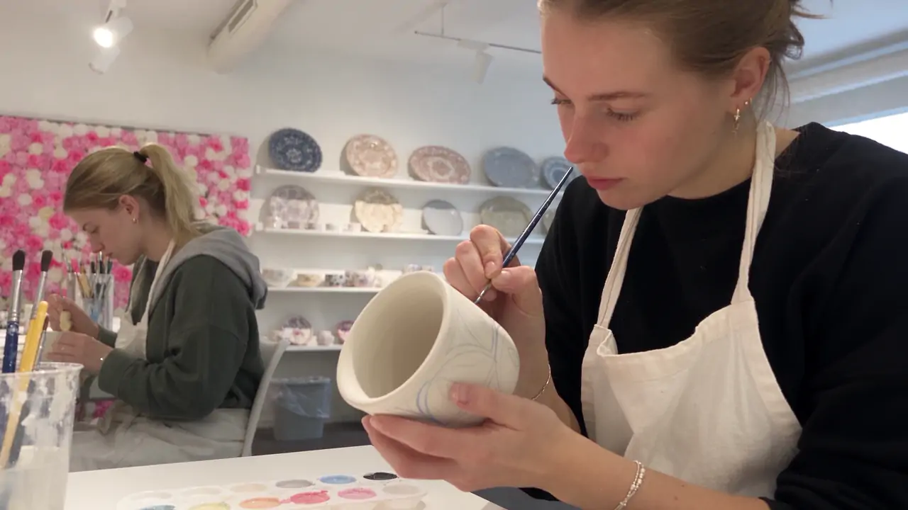 Eine Frau mit weisser Schürze bemalt eine Tasse im Atelier.